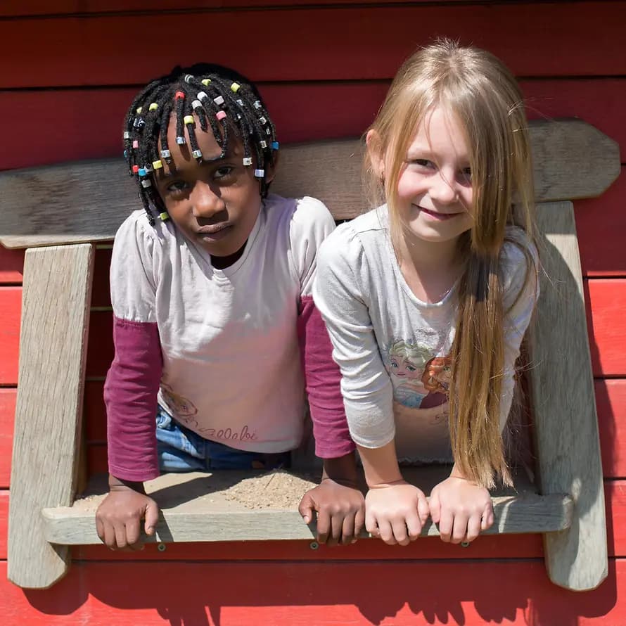 Zwei Kinder lehnen sich lächelnd aus dem Fenster einer Holzhütte.